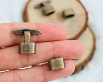 May include: Close-up of several antique bronze-colored metal clasps. One clasp is a T-shape, and another is a rectangular shape. The clasps are on a wooden surface. The clasps are likely for jewelry or other craft projects.