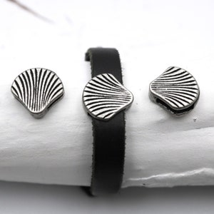 Könnte beinhalten: Drei silberfarbene Metallperlen in Muschelform mit strukturiertem Design. Die Perlen sind auf einem schwarzen Lederband dargestellt.