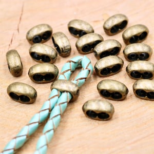 May include: A close-up of a group of antique gold-toned metal beads with two holes each. The beads are arranged on a wooden surface with a turquoise leather cord threaded through one of the beads.