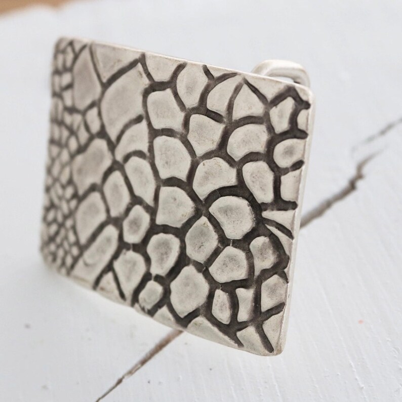 May include: A silver belt buckle with a textured, geometric design. The buckle has a raised, textured pattern that resembles a honeycomb or a web.