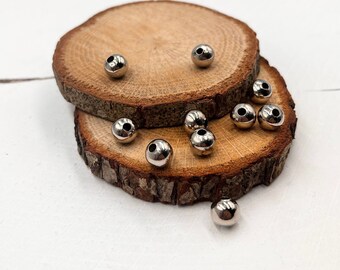 May include: A close-up of a pile of silver metal beads on a wooden surface. The beads are round and have a hole in the center. They are scattered on a wooden surface.
