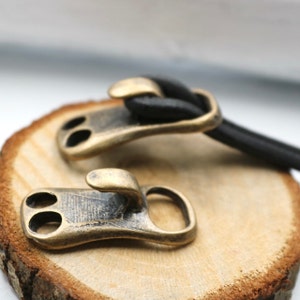 Peut inclure: Deux fermoirs en laiton antique avec un crochet d'un côté et deux trous de l'autre. Les fermoirs sont sur une surface en bois.