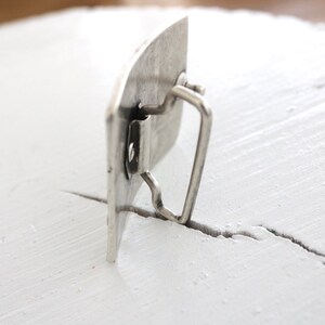 May include: Silver metal belt buckle with a rectangular shape and a smooth, slightly textured surface. The buckle has a simple, functional design with a metal loop for attaching a belt.