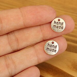 K&ouml;nnte beinhalten: Zwei goldfarbene Metallanh&auml;nger mit der Gravur "hand made". Die Anh&auml;nger sind rund und haben oben ein Loch zum Befestigen an einer Schnur oder einem Band.