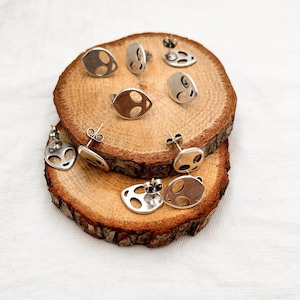 May include: A set of silver alien face earrings on a wooden surface. The earrings are all different shapes and sizes, but they all have the same alien face design.