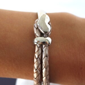 May include: A braided leather bracelet with a silver heart charm and a silver bead. The bracelet is light brown in color.