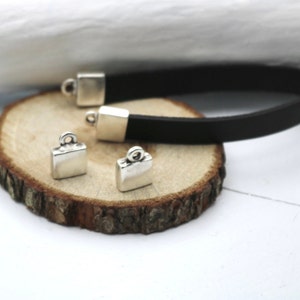Könnte beinhalten: Vier silberfarbene Metall-Anhänger in Form eines kleinen Koffers mit einer Schlaufe oben. Die Anhänger liegen auf einer Holzoberfläche mit einem schwarzen Lederband.