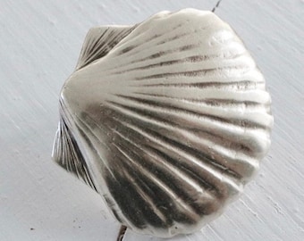 Könnte beinhalten: Ein silberfarbener Muschelschmuck mit einer detaillierten, strukturierten Oberfläche. Die Muschel ist geschlossen und hat eine glatte, abgerundete Form.