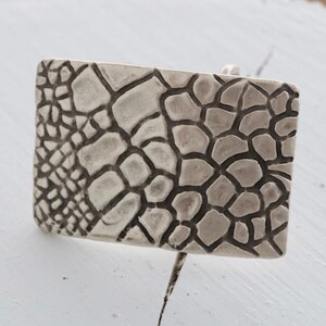 May include: A silver belt buckle with a textured, geometric design resembling a honeycomb or a reptile's skin.