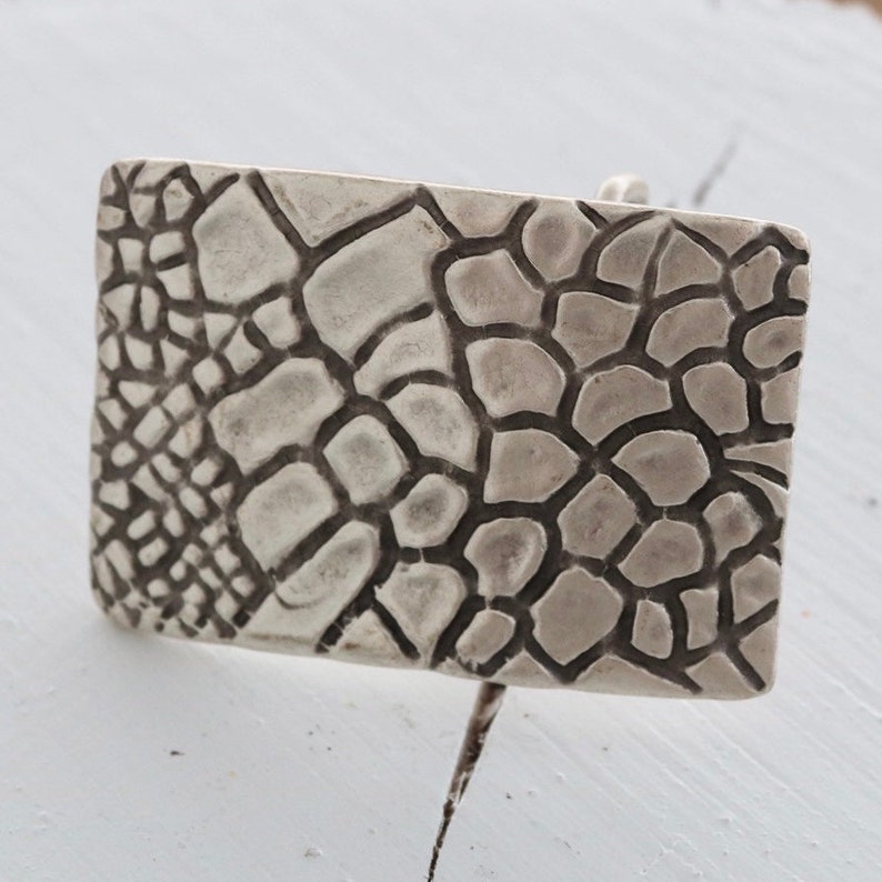 May include: A silver belt buckle with a textured, geometric design resembling a honeycomb or a reptile's skin.