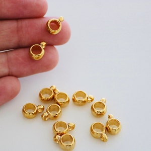 May include: A close-up of ten gold-coloured metal jump rings with a loop on one end. The jump rings are arranged in a semi-circle on a white surface.