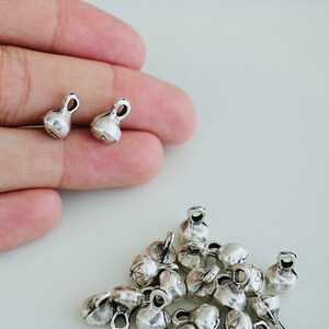 May include: A close-up of silver-toned metal bell charms. The charms are small and have a rounded shape with a loop at the top. There are 17 charms in total, with two charms shown in the hand and the rest in a pile.
