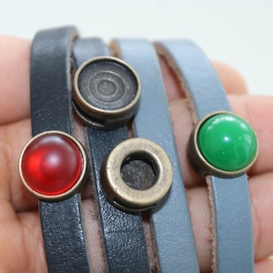 May include: A multi-layered leather bracelet with three different colored metal accents. The accents are a round, bronze-colored piece with a circular design, a round, bronze-colored piece with a hole in the center, and a round, green glass piece.