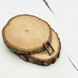 May include: Two round wooden slices with a natural wood grain pattern. A small, rectangular, bronze-coloured metal frame is placed on top of the slices.
