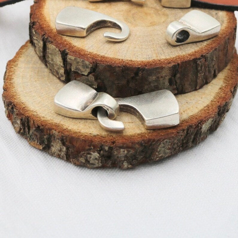 May include: Three silver metal clasps for leather straps. The clasps are shaped like hooks and have a smooth, polished finish.