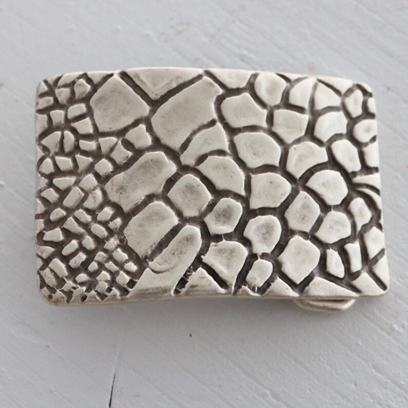 May include: A silver belt buckle with a textured, cracked pattern.