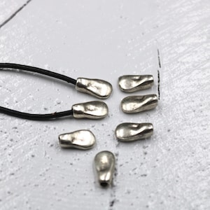 May include: Seven silver-toned metal beads with a slightly irregular, rounded shape. The beads are arranged in a semi-circle on a white surface. A black leather cord is visible in the background.