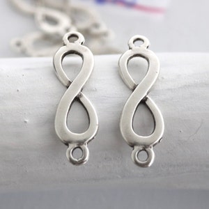 May include: Two silver infinity symbol charms with jump rings at the top and bottom. The charms are on a white surface.