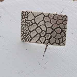 May include: A silver rectangular pendant with a textured, geometric design resembling a honeycomb or a network of veins.