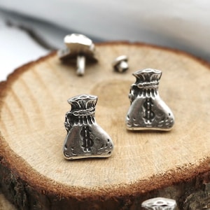 May include: Two silver money bag earrings with a dollar sign on the front. The earrings are on a wooden surface.