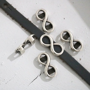 May include: Five silver infinity symbol charms for jewelry making. The charms are on a dark gray leather strap.