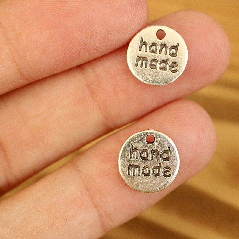 K&ouml;nnte beinhalten: Zwei runde Anh&auml;nger aus silberfarbenem Metall mit der Gravur "hand made".