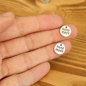May include: Two gold-coloured metal tags with the words "hand made" engraved on them. The tags are round and have a small hole at the top.
