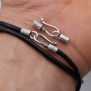May include: A black leather cord bracelet with silver-toned clasps. The bracelet is adjustable and has a simple, minimalist design.