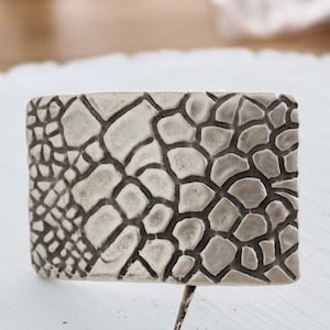 May include: A silver belt buckle with a textured, raised design resembling a reptile skin pattern.