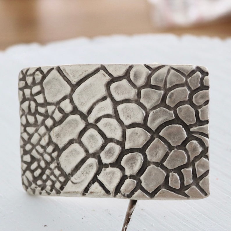 May include: A silver belt buckle with a textured, raised design resembling a reptile skin pattern.