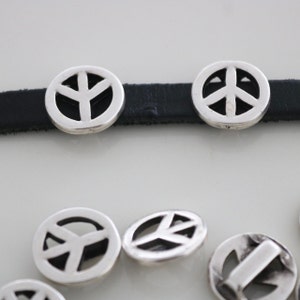 Könnte beinhalten: Silberne Friedenszeichen-Anhänger an einem schwarzen Lederarmband. Die Anhänger sind rund und haben ein Friedenszeichen-Design in der Mitte. Es gibt mehrere Anhänger auf einer weißen Oberfläche.
