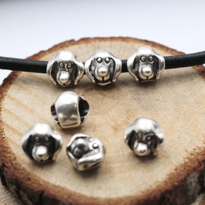 May include: Silver charm beads with a dog face design. The beads are scattered on a wooden surface.