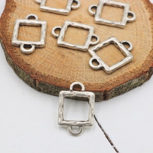Könnte beinhalten: Silberfarbene quadratische Rahmen-Anhänger aus Metall mit gehämmerter Textur. Die Anhänger haben zwei Ösen oben zum Anbringen an Schmuck oder andere Bastelarbeiten.