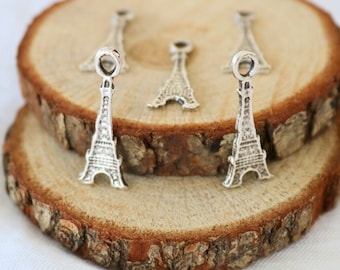 May include: Five silver Eiffel Tower charms on a wooden background. The charms are small and detailed, with a loop at the top for attaching to a necklace or bracelet.