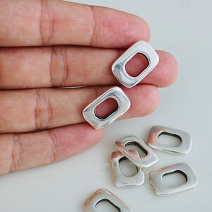 May include: Silver-toned metal jump rings with a rectangular shape and a hole in the center. The rings are arranged in a pile on a white surface.