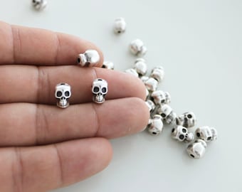 May include: A close-up of a hand holding two silver skull beads. A pile of silver skull beads are scattered on a white surface.
