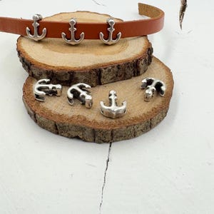May include: Silver anchor charms on a brown leather strap and a wooden surface. The charms are approximately 2.5 cm in size.