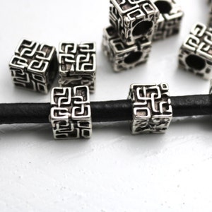May include: Silver metal beads with a geometric design, strung on a black leather cord. The beads are square shaped and have a detailed pattern.