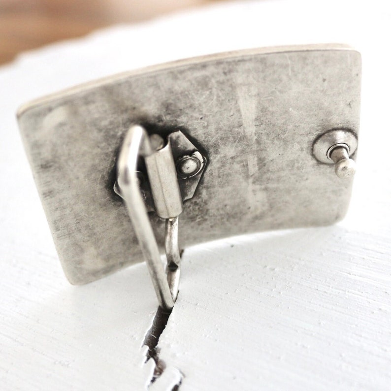 May include: Silver-toned metal belt buckle with a rectangular shape and a textured surface. The buckle has a spring-loaded clasp and a small button on the side.
