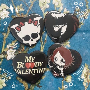 May include: Four heart-shaped pins with black backgrounds and colorful designs. One pin features a skull with pink bows, another has a black and white illustration of a woman with a dark expression, another has a cartoon girl with red hair and a black cat, and the last pin has the text "My Bloody Valentine" with two red hearts.