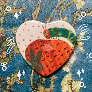 May include: A heart-shaped ceramic pin with a colorful illustration of a caterpillar eating a strawberry. The caterpillar is green with red eyes and a yellow head. The strawberry is red with green leaves.