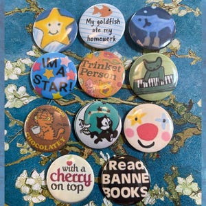 May include: A collection of round pinback buttons with various designs and text. The buttons feature images of a star, a cat playing a keyboard, Garfield, Felix the Cat, and other whimsical illustrations. Text includes "I'M A STAR!", "My goldfish ate my homework", and "Read BANNE BOOKS".