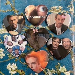 May include: A collection of heart-shaped pinback buttons featuring images of actors and characters. One button has the text "NO VICODIN?" on it. The buttons are in various colors and designs, with a glossy finish.