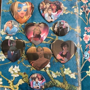 May include: A collection of heart-shaped and round pinback buttons featuring colorful anime-style illustrations. The designs include portraits of various characters with diverse skin tones, clothing, and hairstyles. The buttons are arranged on a blue floral patterned surface.