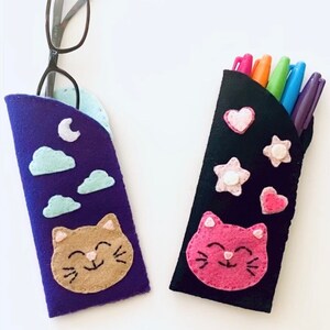 Op de afbeelding: Twee felt pennenkokers met schattige kattengezichten. De ene koker is paars met wolken en een maan, de andere is zwart met roze en witte hartjes en sterren. Beide kokers hebben een glimlachend kattengezicht met roze stiksels.