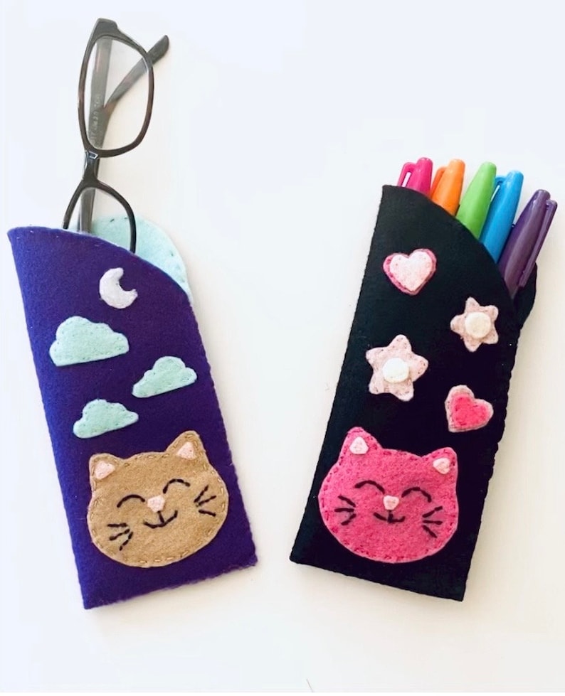 Op de afbeelding: Twee felt pennenkokers met schattige kattengezichten. De ene koker is paars met wolken en een maan, de andere is zwart met roze en witte hartjes en sterren. Beide kokers hebben een glimlachend kattengezicht met roze stiksels.