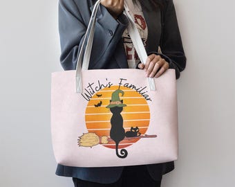May include: A light pink tote bag featuring a black cat wearing a witch's hat, perched on a broomstick in front of an orange sunset. The text "Witch's Familiar" is above the image. White straps and handles.