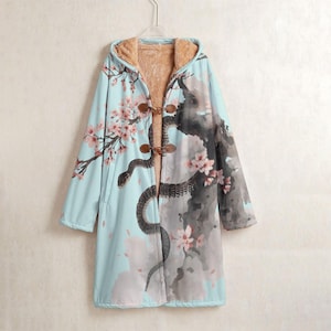 May include: A light blue coat with a hood and toggle fastenings. The coat features a floral and snake design in shades of pink, grey, and black. The interior lining is a tan, fuzzy material. The coat is hanging on a white hanger.