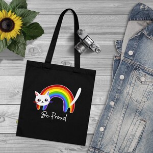 Op de afbeelding: Zwarte tote bag met een witte kat en een regenboog. De kat kijkt naar rechts en de regenboog is erachter. De tekst "Be Proud" staat onder de kat.