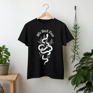 Puede incluir: Camiseta negra con un gráfico blanco de una serpiente envuelta alrededor de una mano que hace el dedo medio. El texto "Bite Back Fear" está escrito en una fuente estilizada sobre la serpiente.